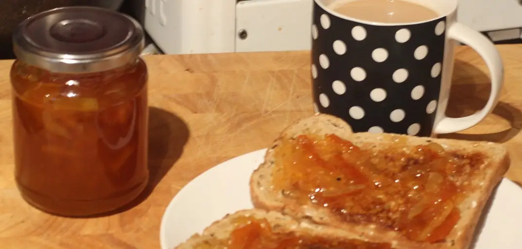 A jar of marmalade, toast and a spotted mug on a wooden board